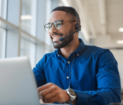 Um homem profissional sorridente, vestindo uma camisa azul e utilizando headset enquanto trabalha em um laptop em um ambiente de escritório. Ele exibe um aparelho ortodôntico, que além de alinhar seus dentes, reflete um impacto positivo na sua autoestima, demonstrando confiança e profissionalismo no local de trabalho.