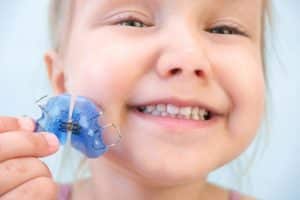 Criança segurando um aparelho móvel infantil de cor azul-esverdeada. O aparelho está destacado na frente de sua boca, evidenciando dentes em fase de troca e sorriso parcial