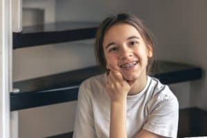 Uma menina sorridente com aparelho ortodôntico, sentada relaxadamente com o queixo apoiado em sua mão, olhando para a câmera. Este retrato ilustra que o uso de aparelho nos dentes pode começar na pré-adolescência, adequado para responder à pergunta "Com quantos anos pode colocar aparelho?"