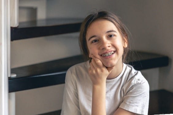 Uma menina sorridente com aparelho ortodôntico, sentada relaxadamente com o queixo apoiado em sua mão, olhando para a câmera. Este retrato ilustra que o uso de aparelho nos dentes pode começar na pré-adolescência, adequado para responder à pergunta "Com quantos anos pode colocar aparelho?