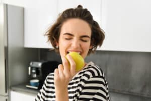 A imagem apresenta uma mulher jovem mordendo uma maçã com entusiasmo e prazer. Ela está em uma cozinha moderna, com um fundo que inclui um armário branco e uma máquina de café. A mulher está vestida com uma blusa de listras horizontais preto e branco, e seu cabelo castanho está preso de forma casual. Sua expressão é animada e sorridente, evidenciando o prazer ao comer a fruta. A fotografia captura um momento dinâmico e alegre, com um foco nítido na maçã e no rosto da mulher, realçando a textura da maçã e a expressão genuína de satisfação.