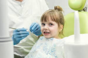 A imagem mostra uma criança no dentista, sendo avaliada para um tratamento ortodôntico preventivo. Ela está sentada confortavelmente em uma cadeira odontológica, vestindo uma bata decorada com personagens de desenho animado, o que contribui para uma atmosfera amigável e acolhedora para crianças. O dentista, de luvas azuis, prepara-se para examinar a boca da menina com instrumentos dentários, indicando uma checagem cuidadosa que é parte do processo de avaliação ortodôntica. A criança parece relaxada e um pouco curiosa sobre o procedimento, mostrando confiança e tranquilidade durante a consulta.