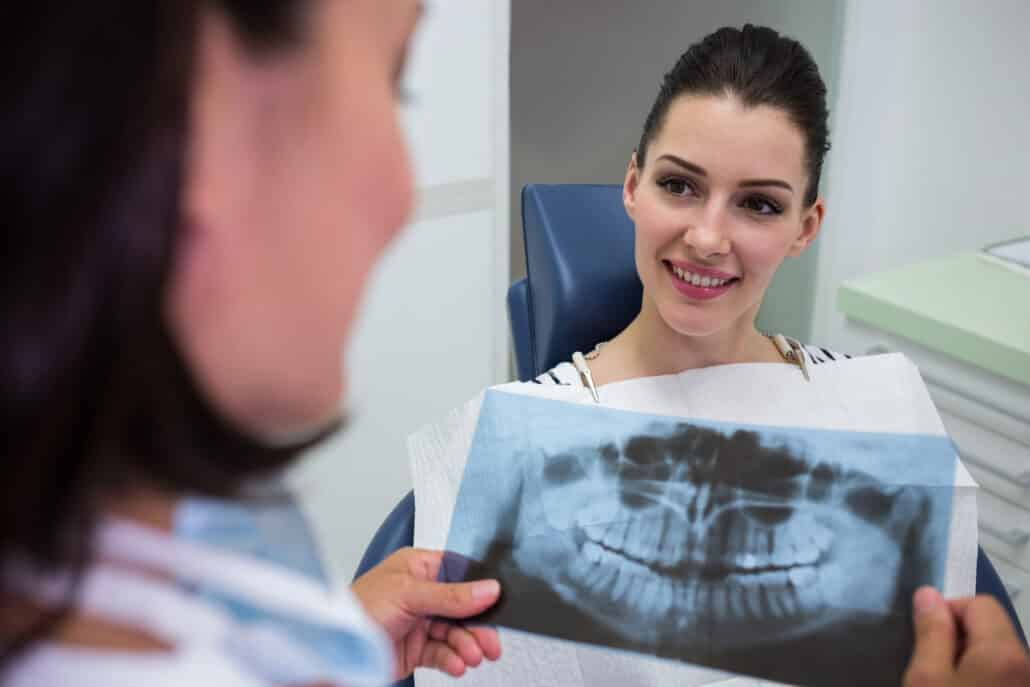 uma paciente mulher está sorrindo enquanto examina uma radiografia de sua mandíbula que lhe é mostrada por sua dentista, que está parcialmente visível. A paciente, com cabelo puxado para trás e uma expressão alegre, está sentada em uma cadeira odontológica em um consultório claro e moderno. A dentista, que aparece de perfil, está discutindo as nuances da radiografia, possivelmente como parte de uma consulta para documentação ortodôntica ou diagnóstico. 