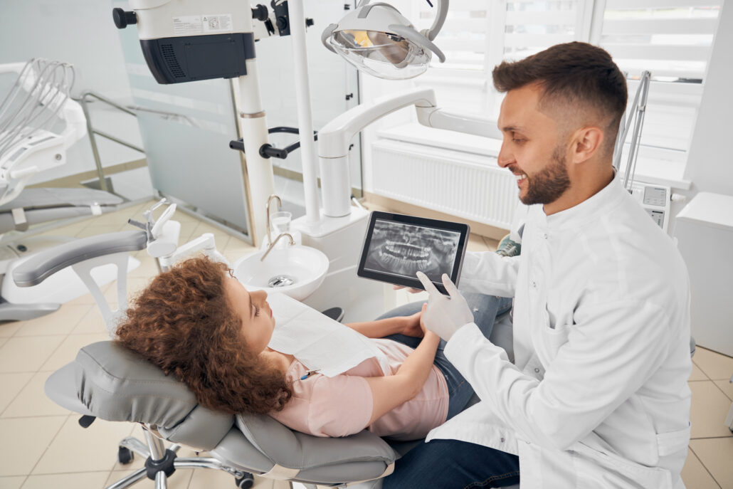 dentista masculino, vestido com jaleco branco, mostrando uma radiografia dentária em um tablet para uma paciente jovem, que está reclinada em uma cadeira odontológica. Eles estão em um consultório dentário moderno e bem iluminado. A paciente, que tem cabelos cacheados castanhos e veste uma blusa rosa, olha atentamente para o tablet, sugerindo que estão discutindo sua documentação ortodôntica. 