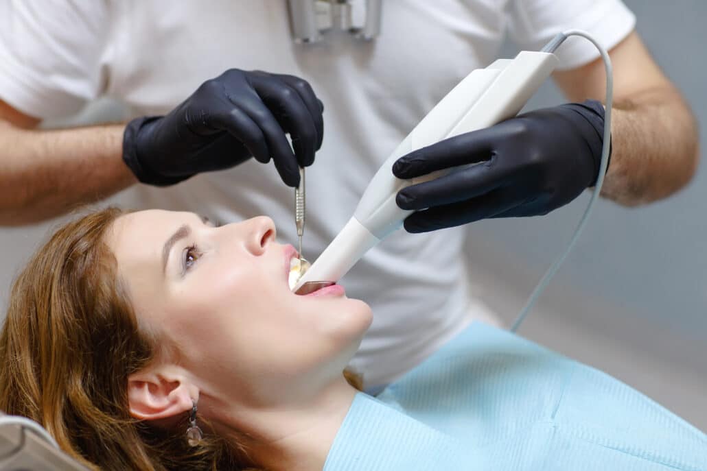 ma paciente mulher está recebendo atendimento odontológico em uma cadeira de dentista. Um dentista masculino, usando luvas pretas e camiseta branca, está realizando um procedimento com um scanner intraoral 3D, que é utilizado para capturar imagens digitais precisas dos dentes da paciente. A paciente, de cabelos castanhos e com uma expressão tranquila, parece estar colaborando durante o procedimento.