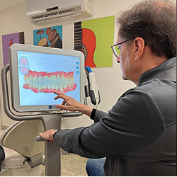 Ortodontia antes e depois: Dr. Pedro Barini, um especialista em ortodontia, está usando um software de simulação para planejar tratamentos ortodônticos. A imagem mostra ele apontando para um modelo 3D colorido dos dentes no monitor, demonstrando as etapas previstas para o alinhamento dental do paciente.