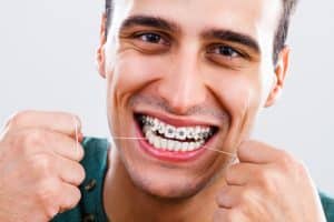 Homem jovem utilizando fio dental enquanto sorri, mostrando aparelho ortodôntico metálico nos dentes. 