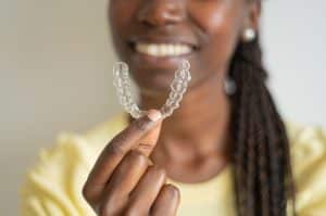 Mulher sorridente de pele escura, vestindo uma blusa amarela e segurando um alinhador invisível com uma das mãos. Ela tem cabelos longos e trançados, e o foco está no alinhador transparente que ela exibe de perto.